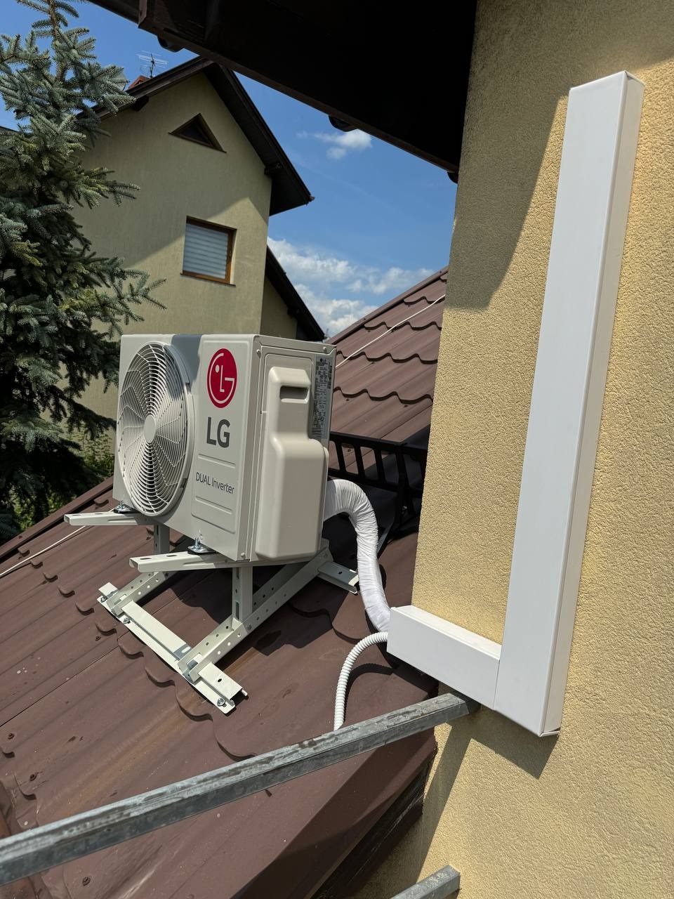 Zewnętrzna jednostka klimatyzacji LG Dual Inverter zamontowana na metalowej konstrukcji na dachu, z widoczną białą osłoną rury przy elewacji budynku.