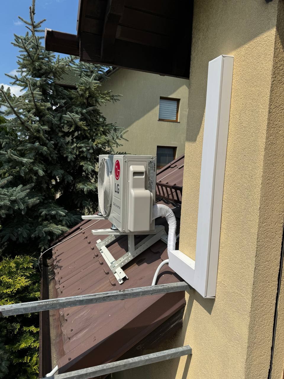 Zewnętrzna jednostka klimatyzacji LG Dual Inverter zamontowana na wsporniku na dachu domu, z widoczną osłoną instalacji na elewacji budynku.