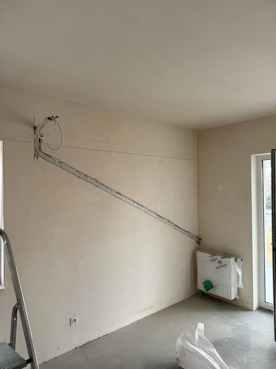 Wnętrze w trakcie prac instalacyjnych: rury poprowadzone po ścianie, nowy grzejnik zabezpieczony folią, widoczna drabina malarska i ślady po kuciu w ścianie.