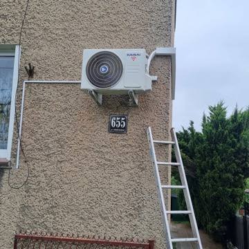 Biała jednostka zewnętrzna klimatyzacji marki Kaisai zamontowana na ścianie budynku obok numeru domu 655. Widoczna drabina oparta o ścianę, sugerująca świeży montaż.