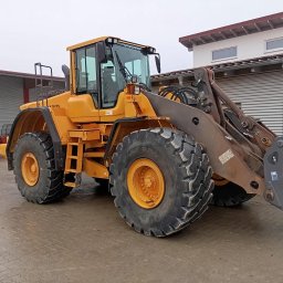 TRANSBUD SILESIA SPÓŁKA Z OGRANICZONĄ ODPOWIEDZIALNOŚCIĄ - Żółta ładowarka kołowa Volvo z widocznymi śladami użytkowania i błotem na oponach, zaparkowana na placu z budynkami w tle.