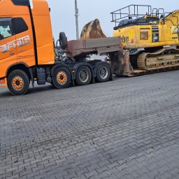 TRANSBUD SILESIA SPÓŁKA Z OGRANICZONĄ ODPOWIEDZIALNOŚCIĄ - Pomarańczowa ciężarówka z naczepą transportuje żółtą koparkę Komatsu PC 360 LC na placu wyłożonym kostką brukową.