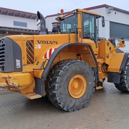 TRANSBUD SILESIA SPÓŁKA Z OGRANICZONĄ ODPOWIEDZIALNOŚCIĄ - Żółta ładowarka kołowa Volvo L180F, widok z boku, na tle budynku z szarymi oknami i bramą garażową.