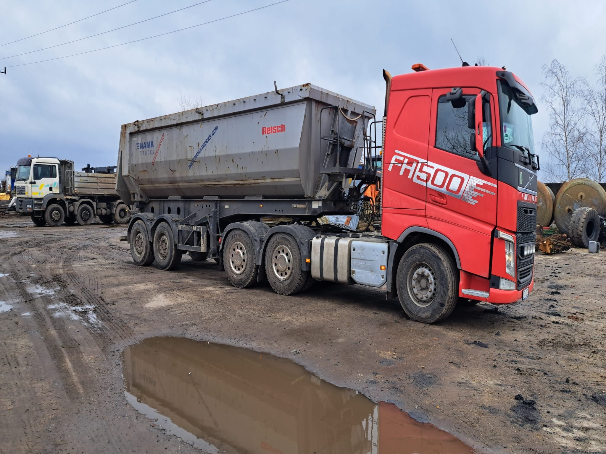 Czerwona ciężarówka Volvo FH500 z naczepą wywrotką Reisch, zaparkowana na błotnistym terenie, w tle inne pojazdy i opony.