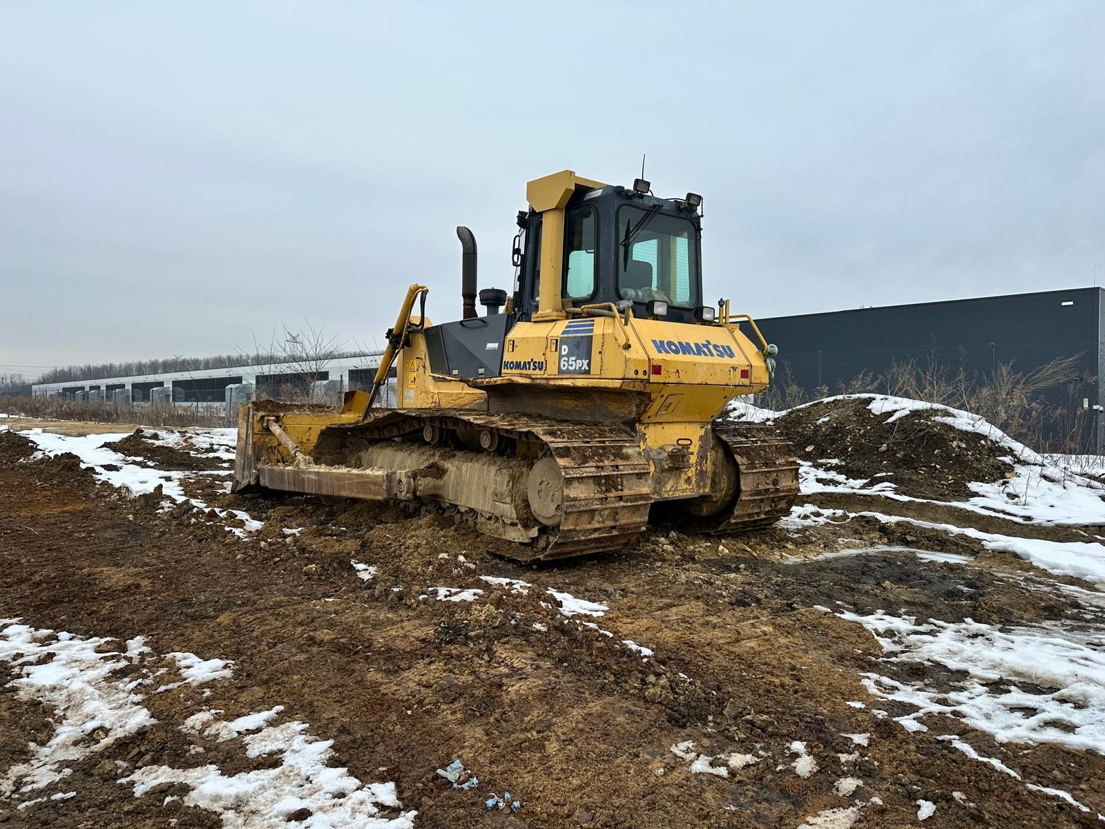 Żółty spychacz Komatsu 65PX w Tarnowskich Górach stoi na placu budowy z resztkami śniegu, przygotowany do niwelacji terenu pod budowę hali.