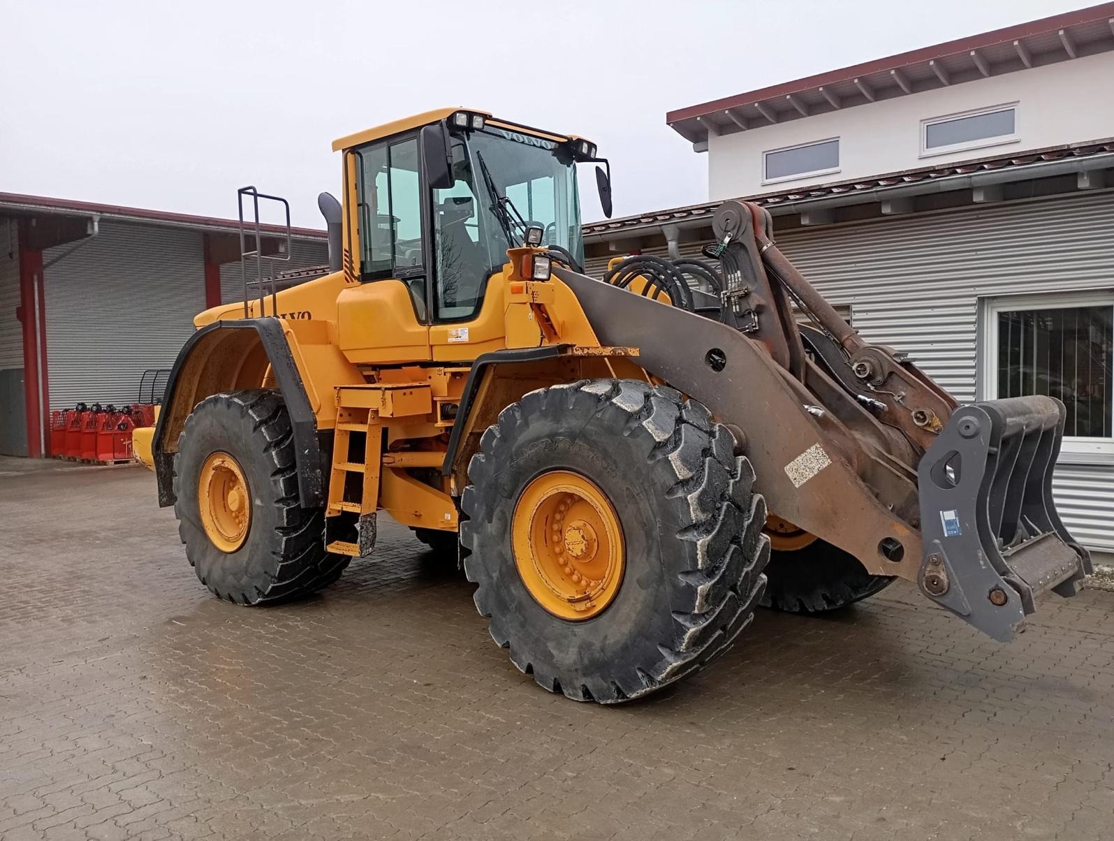 Żółta ładowarka kołowa Volvo z widocznymi śladami użytkowania i błotem na oponach, zaparkowana na placu z budynkami w tle.