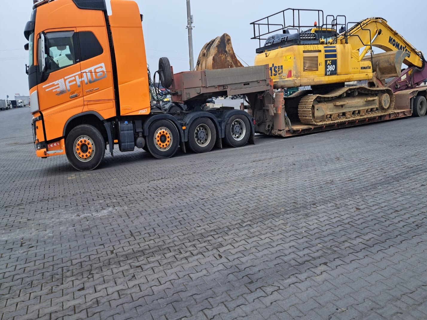 Pomarańczowa ciężarówka z naczepą transportuje żółtą koparkę Komatsu PC 360 LC na placu wyłożonym kostką brukową.