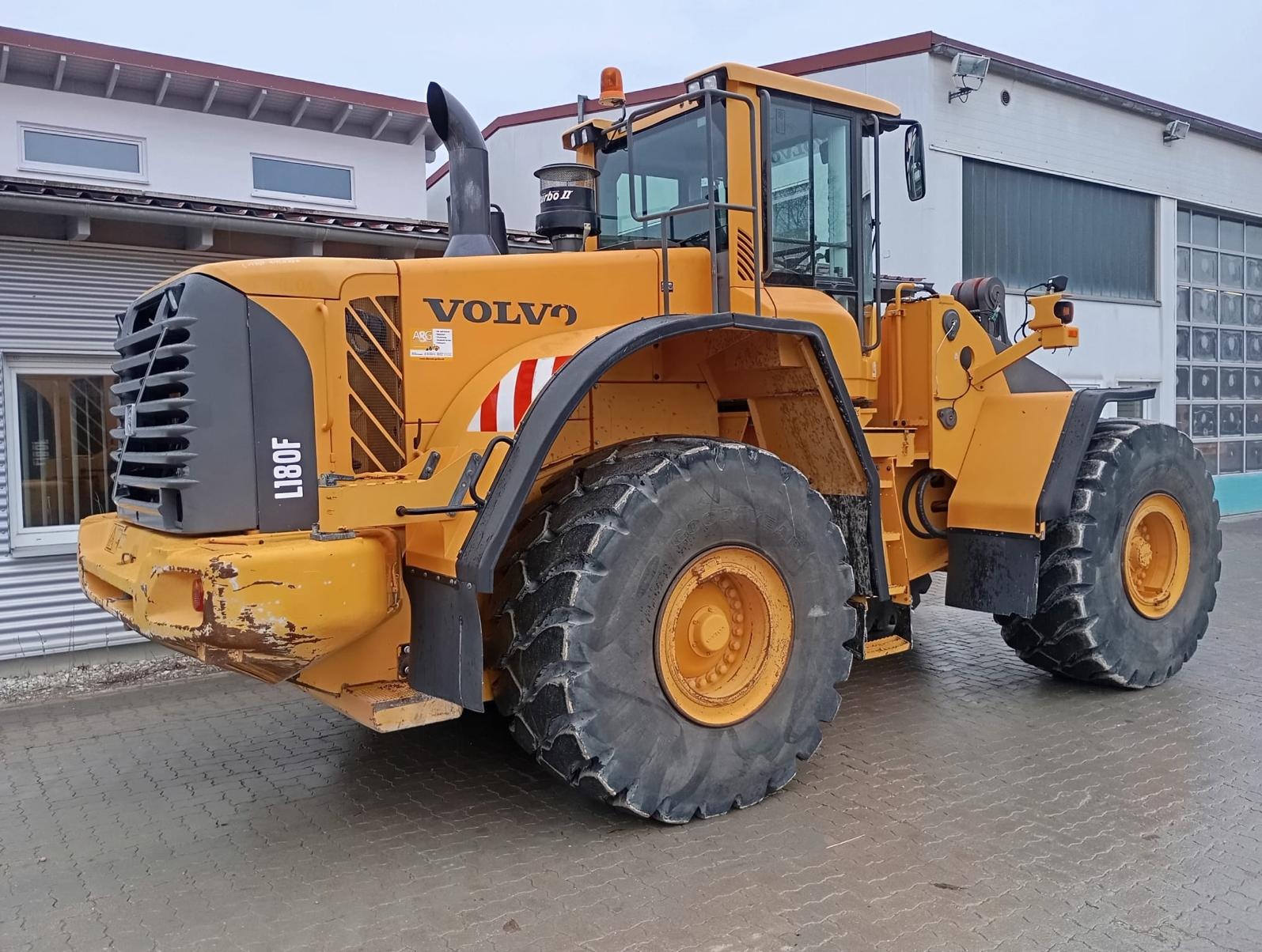 Żółta ładowarka kołowa Volvo L180F, widok z boku, na tle budynku z szarymi oknami i bramą garażową.