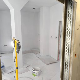 Luckybud - Wnętrze po malowaniu na biało, widoczne przygotowania do dalszych prac wykończeniowych. Na pierwszym planie żółty statyw z lampą i wiadro z farbą.