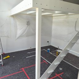 Luckybud - Biała konstrukcja antresoli z drabinką w trakcie malowania. Podłoga zabezpieczona, ściany częściowo zakryte folią malarską. Widoczne narzędzia malarskie.