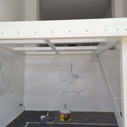 Luckybud - Biała konstrukcja antresoli w trakcie malowania, zabezpieczona folią malarską. Widoczne wiadro z farbą i wałek. Minimalistyczne wnętrze w trakcie prac wykończeniowych.