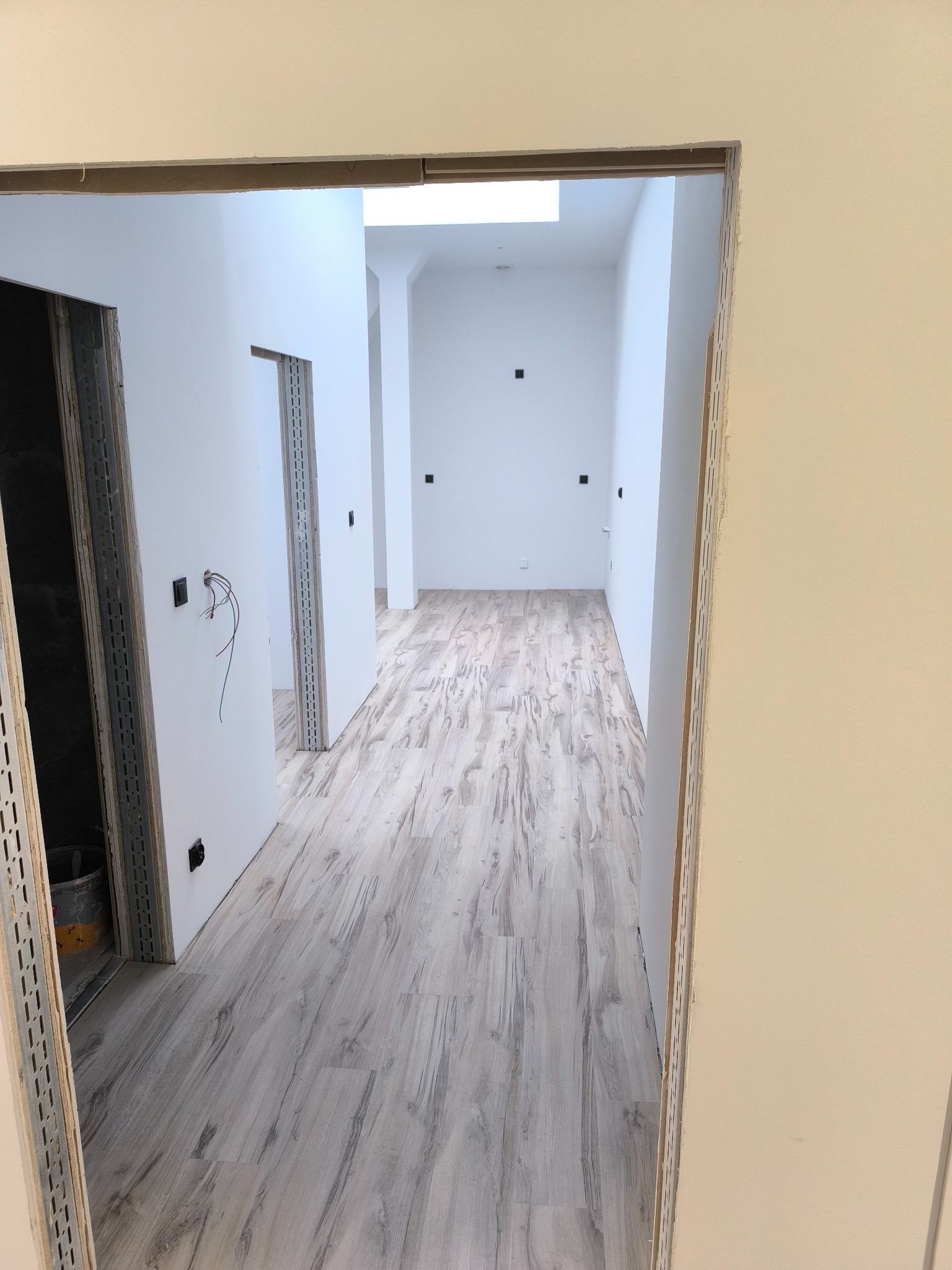 Korytarz w trakcie wykańczania: ułożone panele podłogowe, ściany przygotowane do malowania, widoczne kable elektryczne. Ujęcie z perspektywy otwartych drzwi.