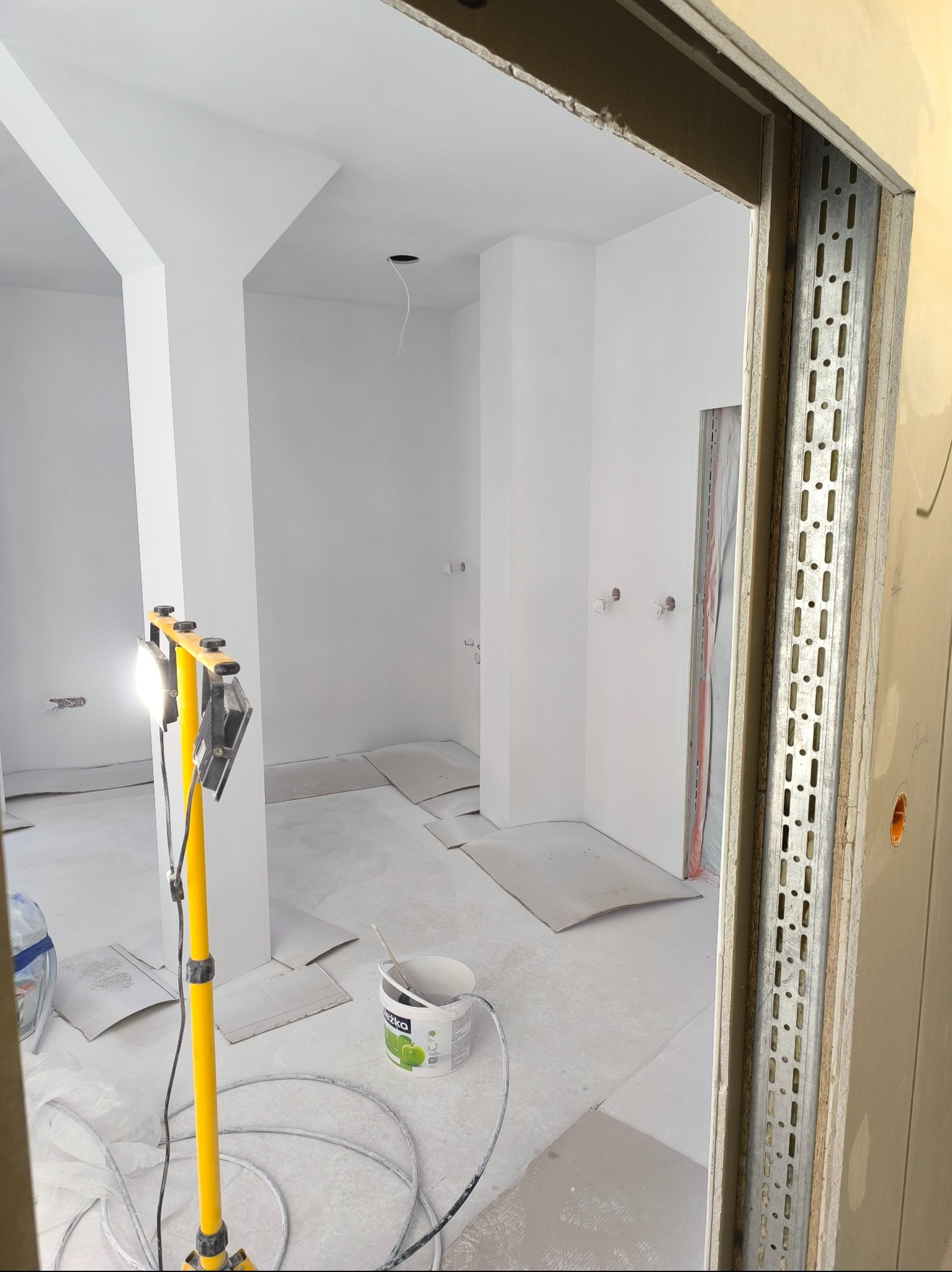 Wnętrze po malowaniu na biało, widoczne przygotowania do dalszych prac wykończeniowych. Na pierwszym planie żółty statyw z lampą i wiadro z farbą.