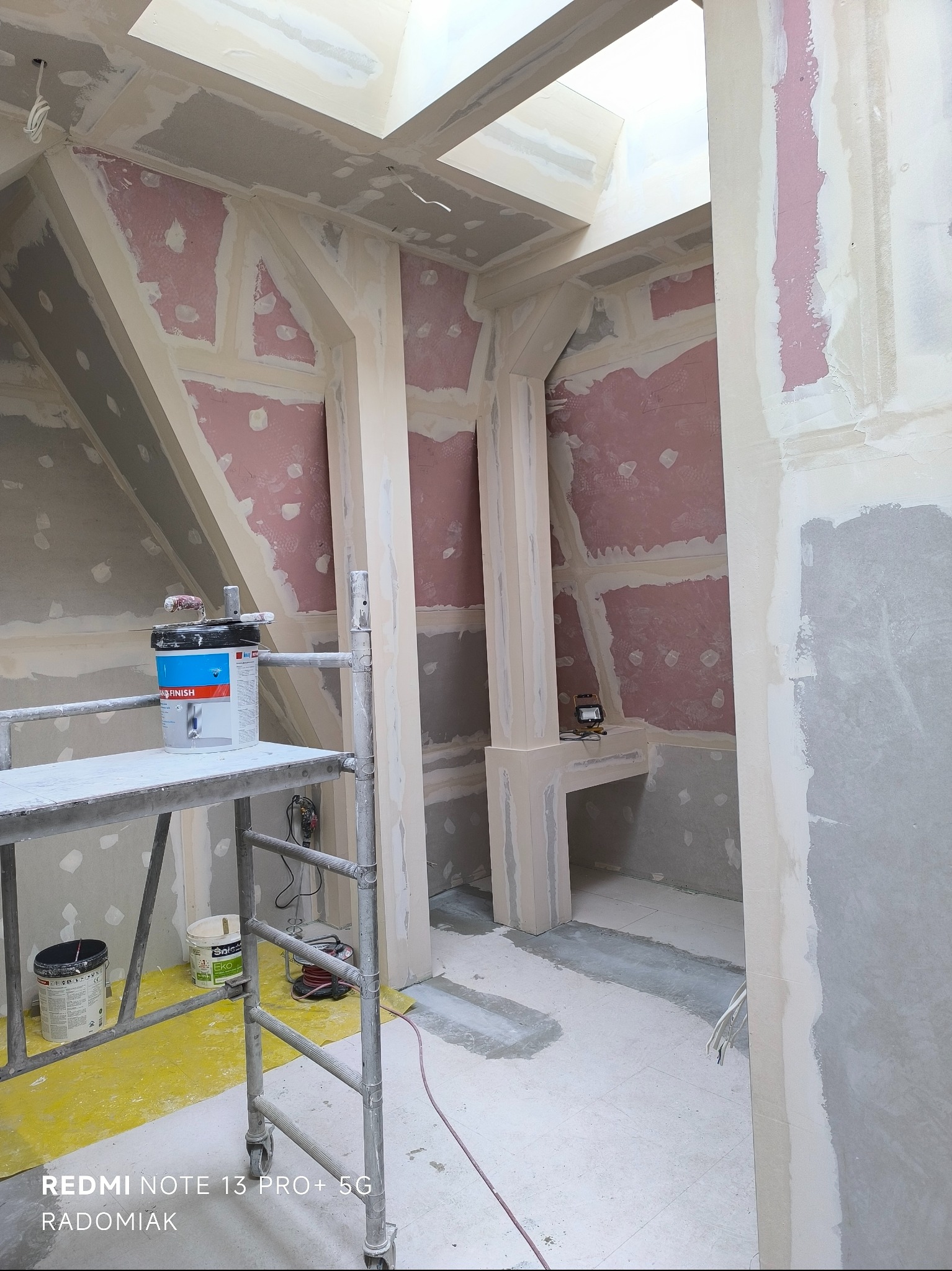 Skośne poddasze w trakcie szpachlowania: widoczne płyty gipsowe, rusztowanie z farbą i narzędziami. Oświetlenie naturalne z okna dachowego.