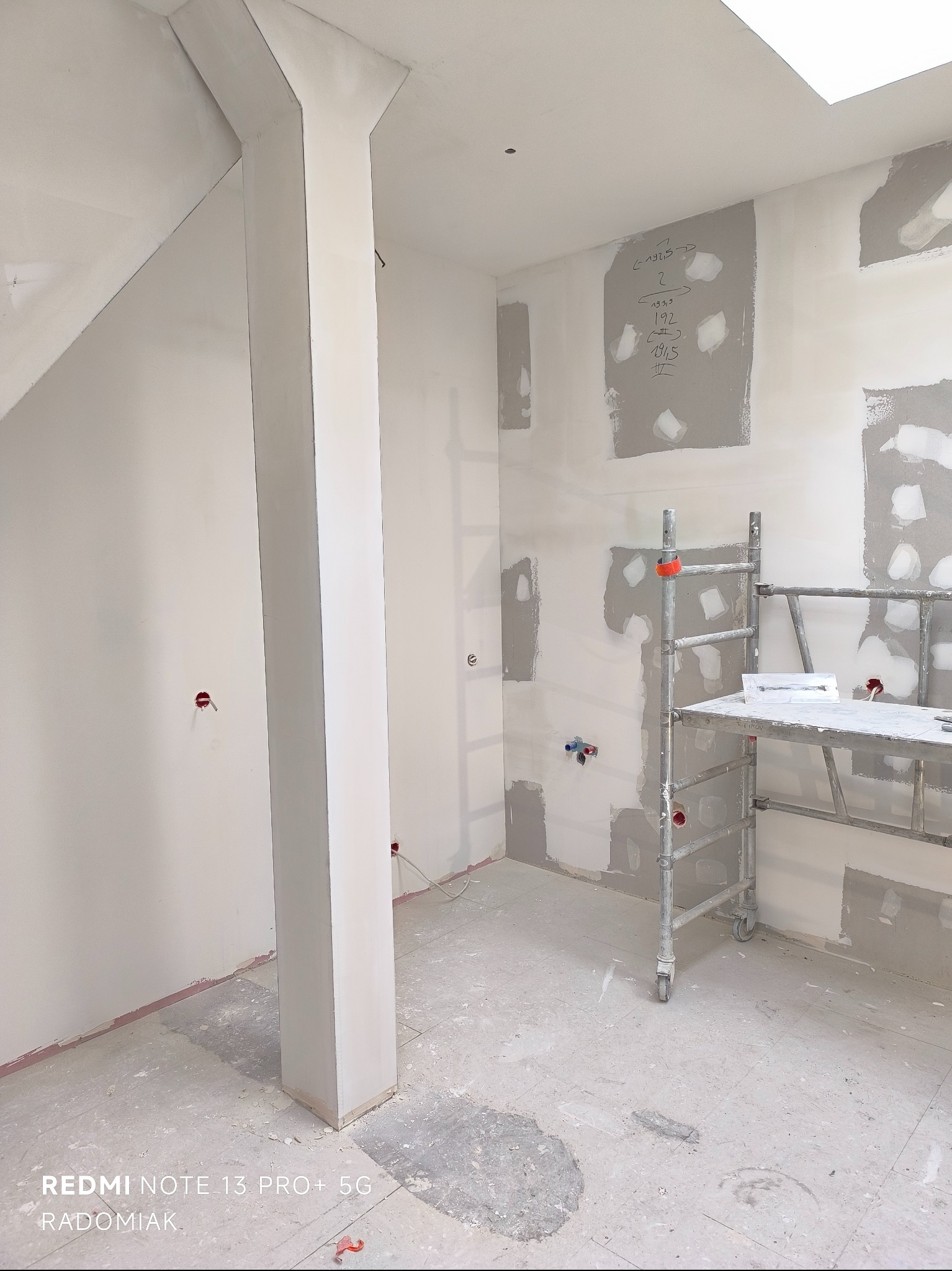 Wnętrze w trakcie szpachlowania: ściany z łatami szpachli, aluminiowe rusztowanie, kolumna obłożona płytą gipsową, sufit z oknem dachowym. Widoczne ślady po pracach.