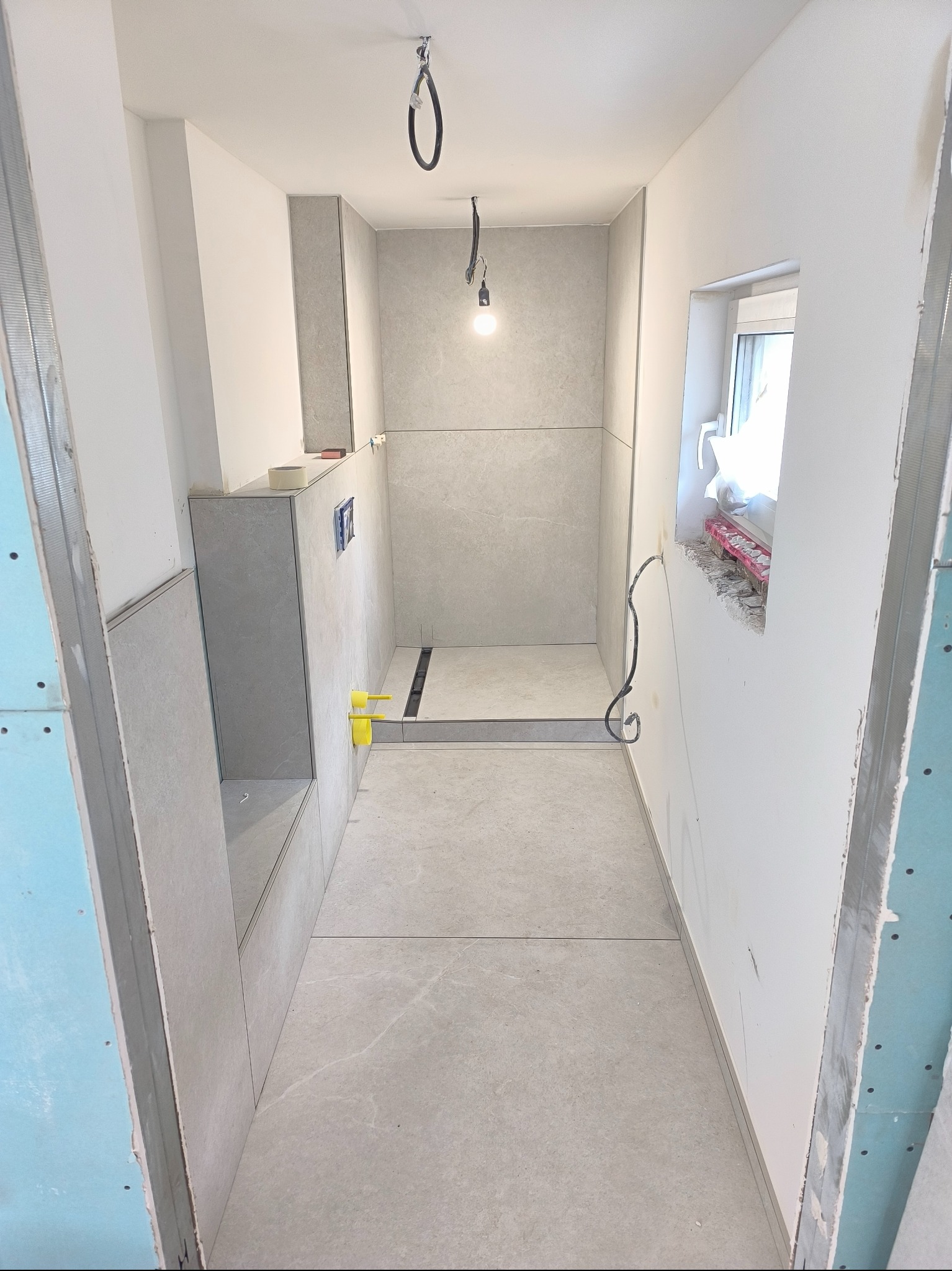 Wnętrze łazienki w trakcie wykańczania: szare płytki, widoczny odpływ liniowy, instalacja elektryczna przy oknie, płyty karton-gips na ścianach. Surowy stan.