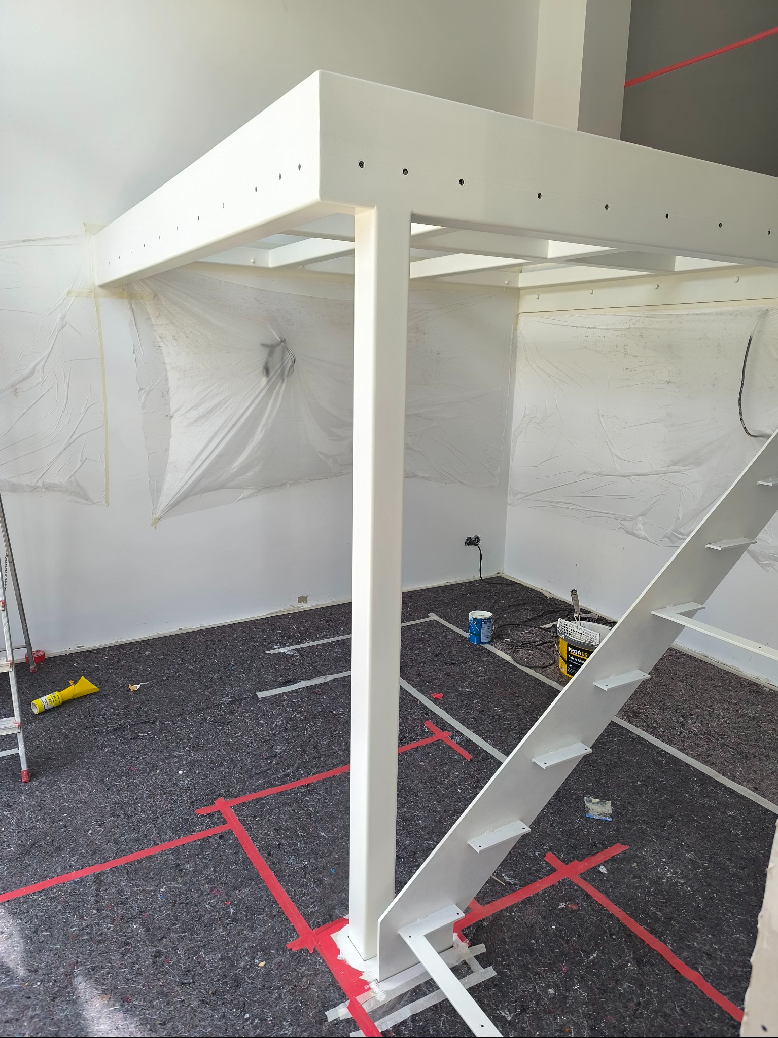 Biała konstrukcja antresoli z drabinką w trakcie malowania. Podłoga zabezpieczona, ściany częściowo zakryte folią malarską. Widoczne narzędzia malarskie.