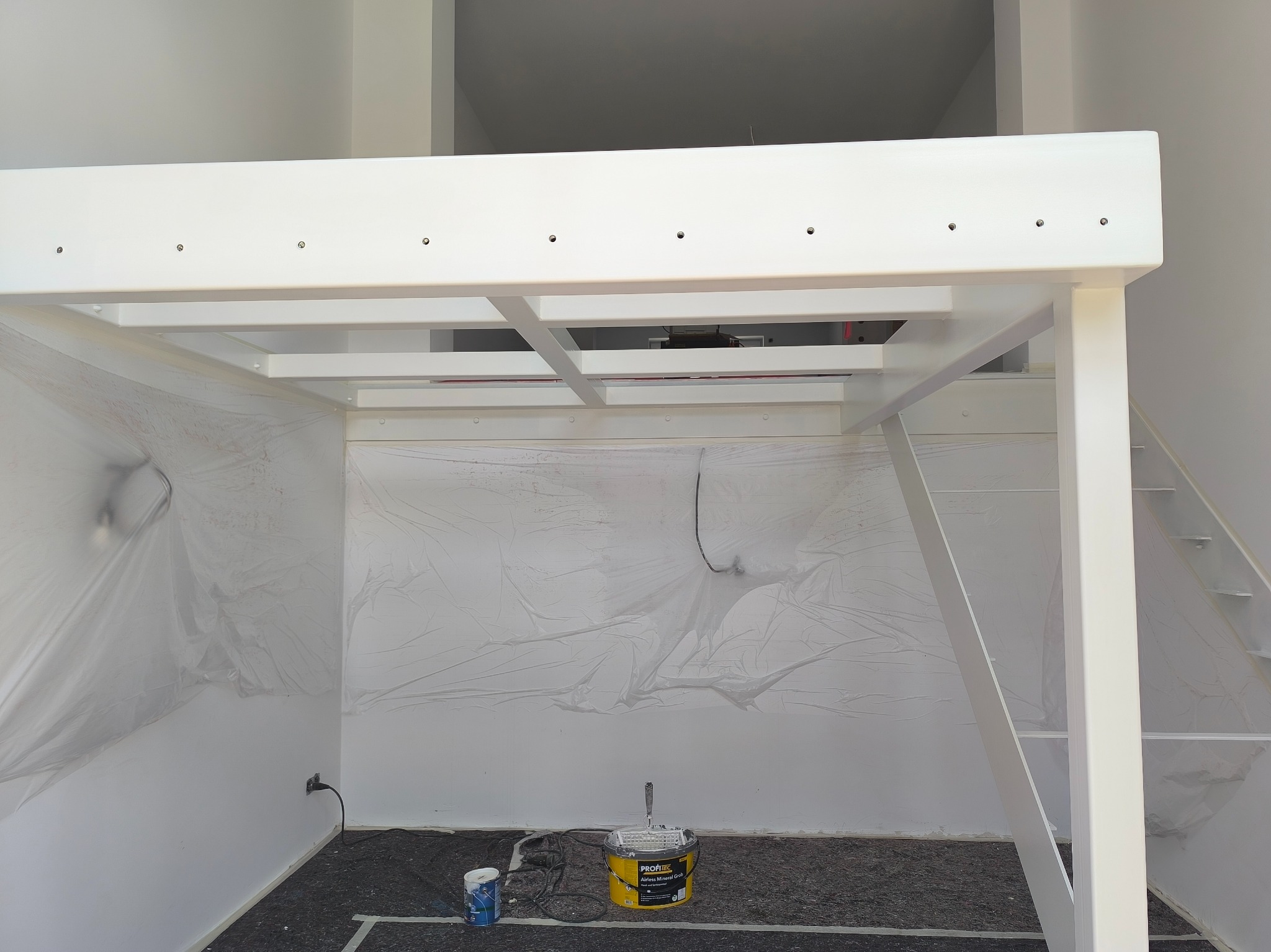 Biała konstrukcja antresoli w trakcie malowania, zabezpieczona folią malarską. Widoczne wiadro z farbą i wałek. Minimalistyczne wnętrze w trakcie prac wykończeniowych.