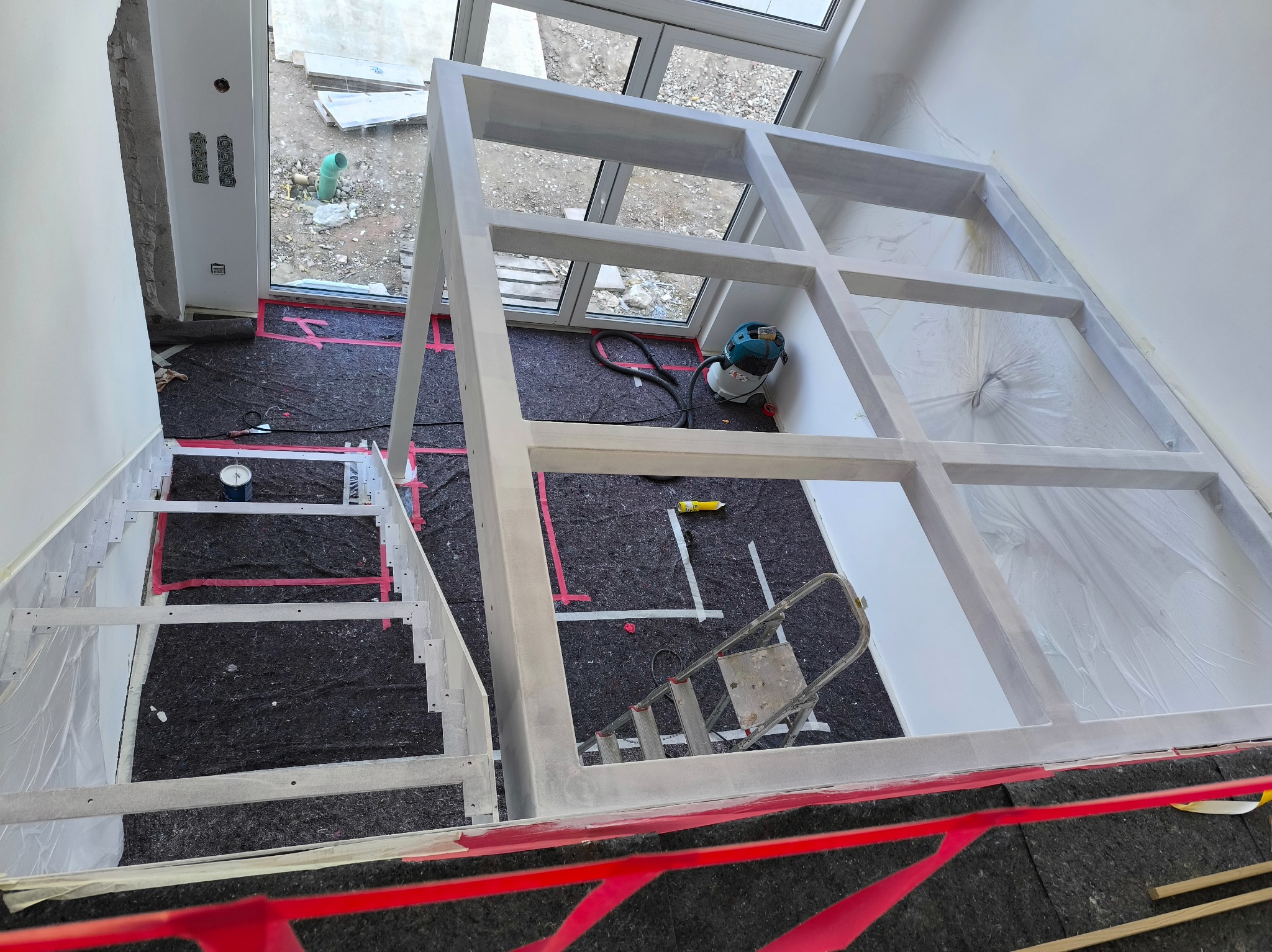 Biała konstrukcja szkieletowa poddasza z oknami, zabezpieczona folią malarską, widok z góry. Na podłodze mata ochronna i drabina. Etap prac wykończeniowych.