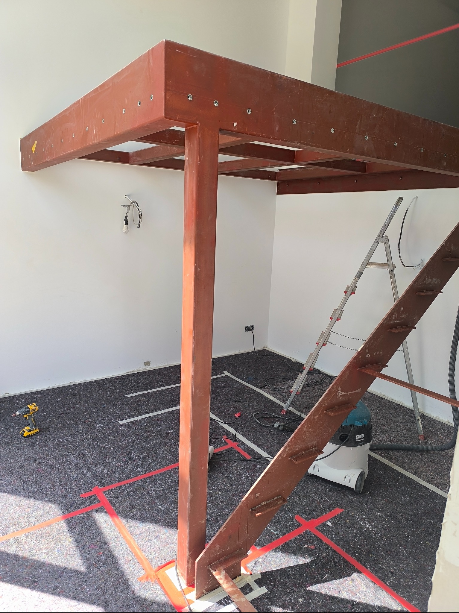 Stalowa konstrukcja antresoli w trakcie montażu, widoczne narzędzia i drabina. Podłoga zabezpieczona, wyznaczona laserem. Oświetlenie surowe, wnętrze w trakcie prac.