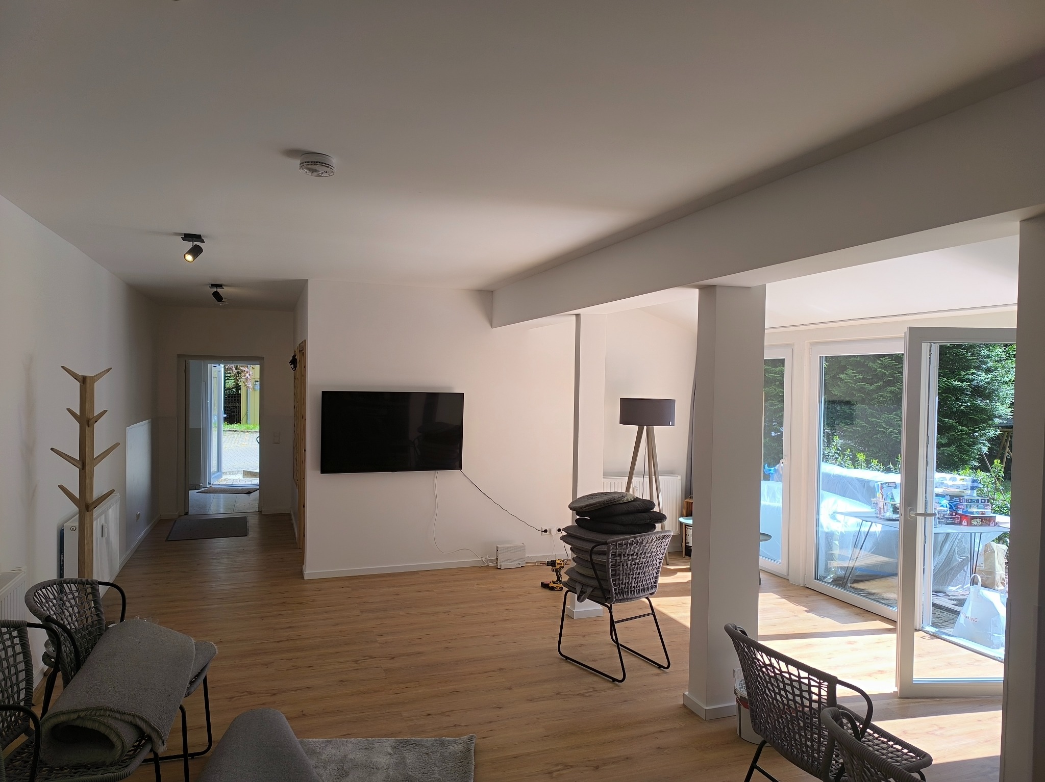 Jasne wnętrze po remoncie: drewniana podłoga, białe ściany, nowoczesne meble i duże okna tarasowe wpuszczające dużo światła dziennego. Widoczny fragment sofy i krzesła.