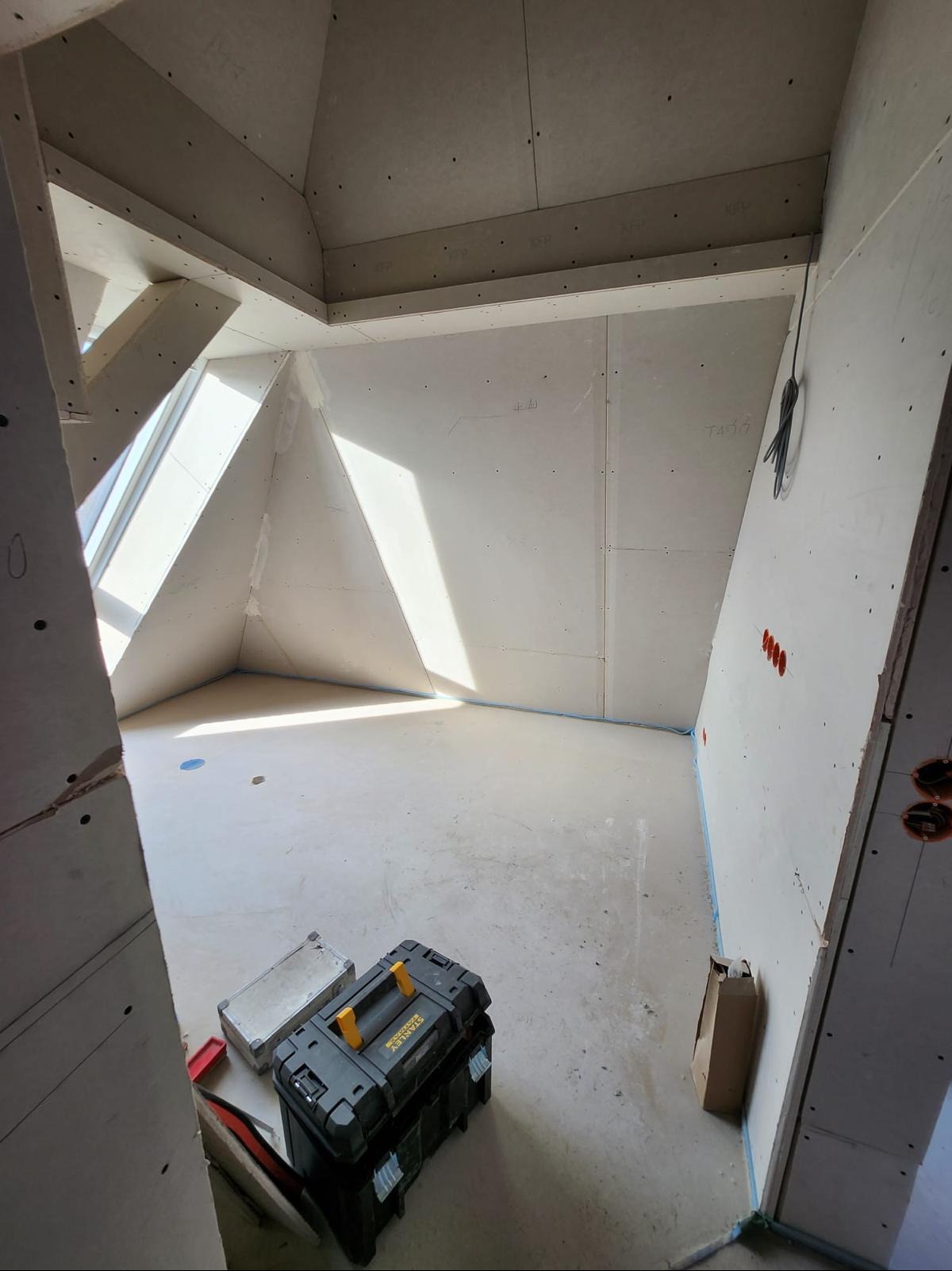 Pokój ze skośnym oknem dachowym w trakcie montażu płyt karton-gips, widoczne narzędzia i kable elektryczne. Ściany przygotowane do dalszych prac wykończeniowych.