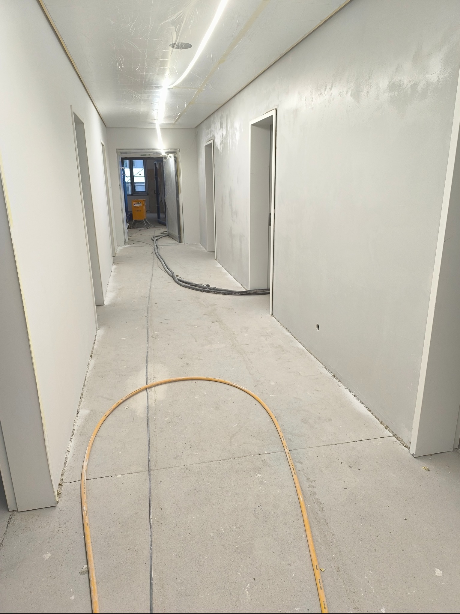 Korytarz w trakcie szpachlowania ścian na gładko, zabezpieczony folią sufit, widoczne węże i sprzęt do szpachlowania. Ściany w jasnym odcieniu szarości.