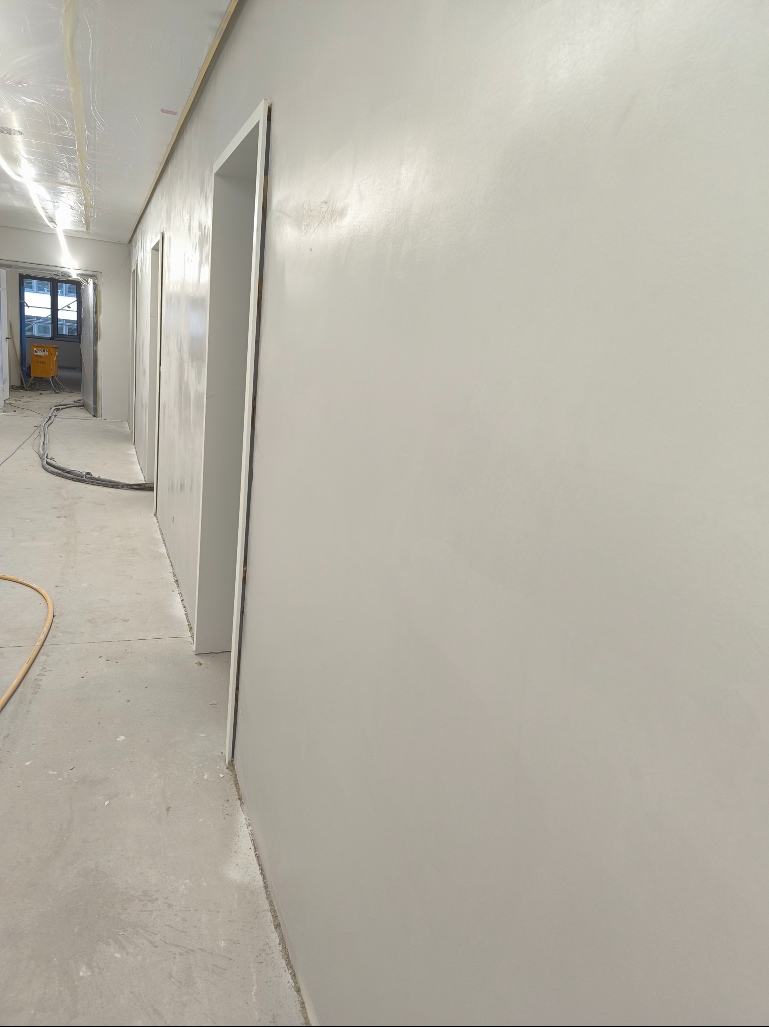 Gładka, biała ściana po szpachlowaniu w jasnym wnętrzu, widoczne ościeżnice drzwiowe i sprzęt budowlany w tle. Minimalistyczny kadr, świeżo wykończone powierzchnie.