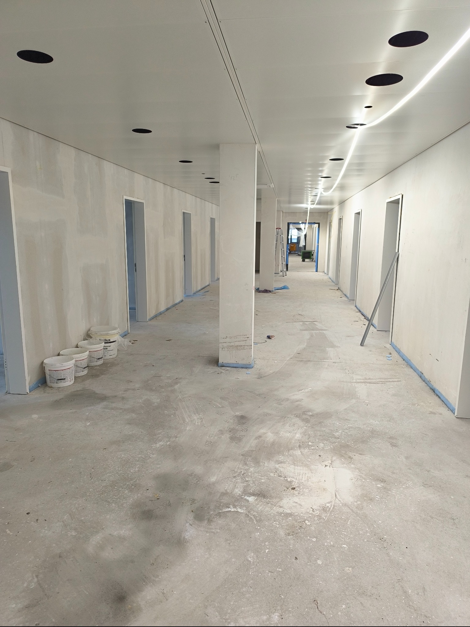 Korytarz biurowy w trakcie szpachlowania i malowania, widoczne puszki z farbą, oświetlenie sufitowe i przygotowane ściany. Długi kadr z perspektywą.