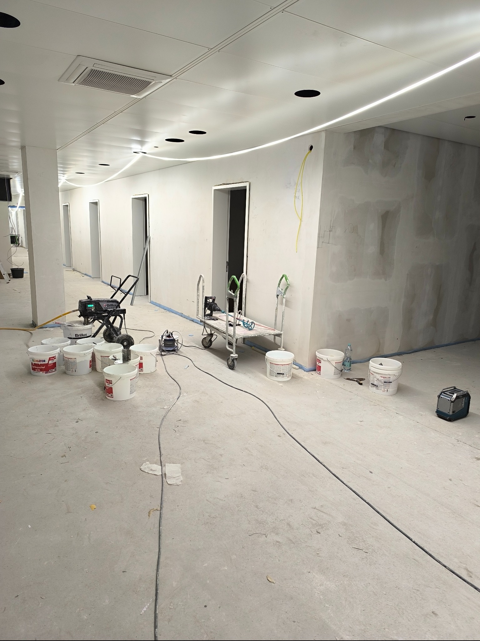 Wnętrze w trakcie szpachlowania: ściany pokryte gładzią, sprzęt do natrysku, wiadra z masą szpachlową, w tle widoczne otwory drzwiowe i oświetlenie liniowe.
