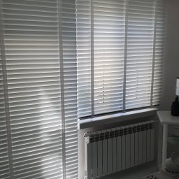 SIGMA SYSTEMY OSŁONOWE TOMASZ ŚLEMP - Białe żaluzje drewniane poziome, częściowo zasłonięte, wpadające światło słoneczne, na tle grzejnik i fragment stolika. Nowoczesny design wnętrza.