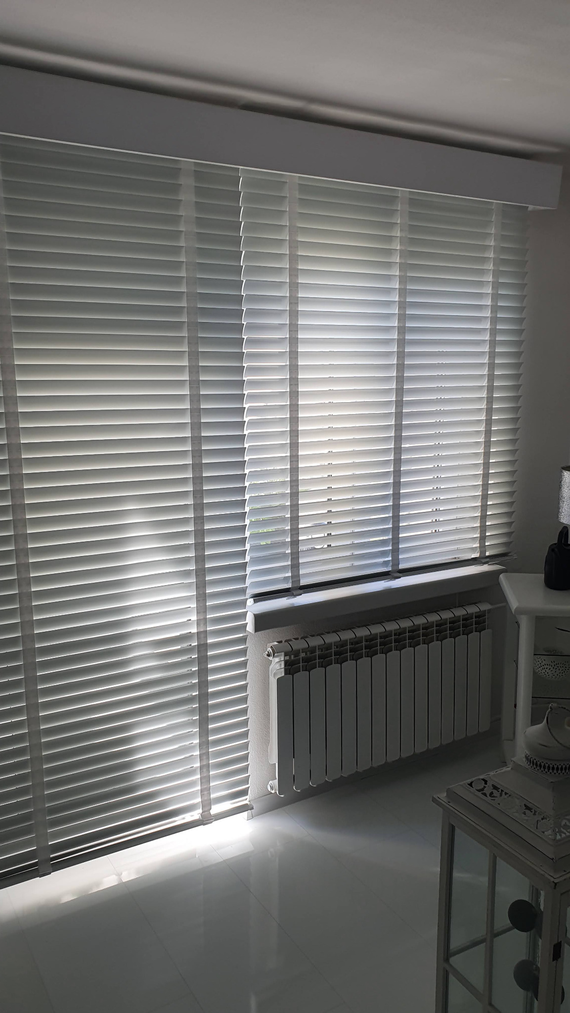 Białe żaluzje drewniane poziome, częściowo zasłonięte, wpadające światło słoneczne, na tle grzejnik i fragment stolika. Nowoczesny design wnętrza.
