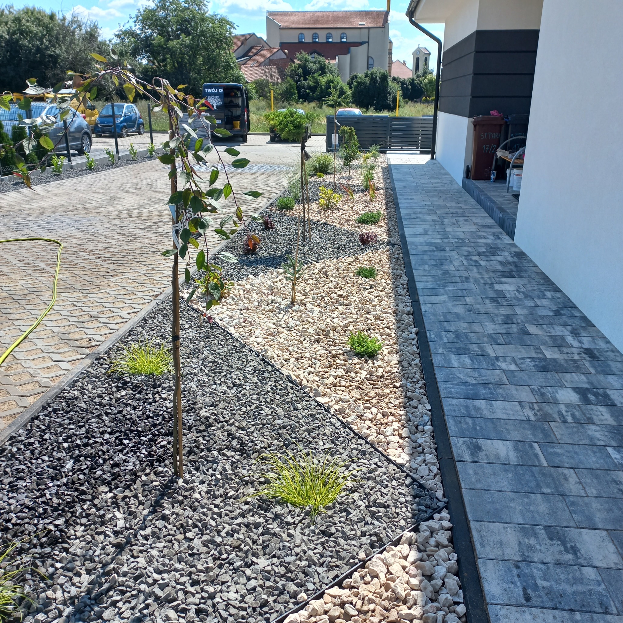 Nowoczesny ogród przydomowy z geometrycznym podziałem na strefy: ciemny żwir, jasne kamienie i rośliny ozdobne, obok ścieżka z szarych płyt chodnikowych.