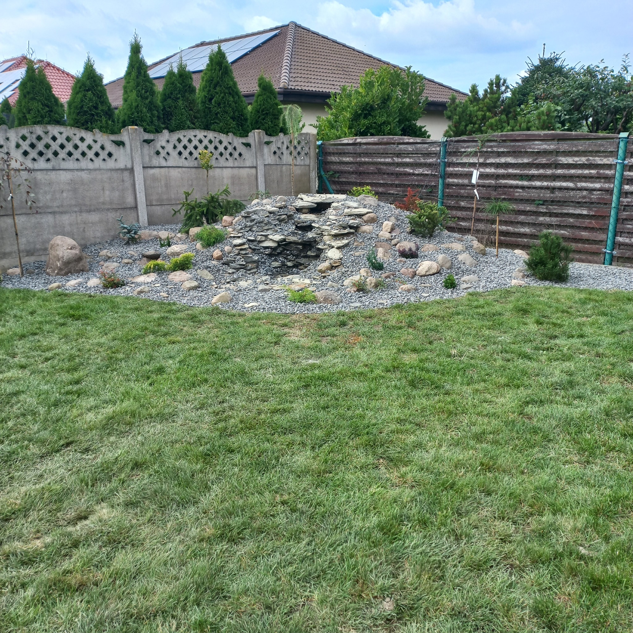 Ogród z kaskadą wodną zbudowaną z płaskich kamieni, otoczoną ozdobnymi kamykami i niską roślinnością, widok z perspektywy trawnika.