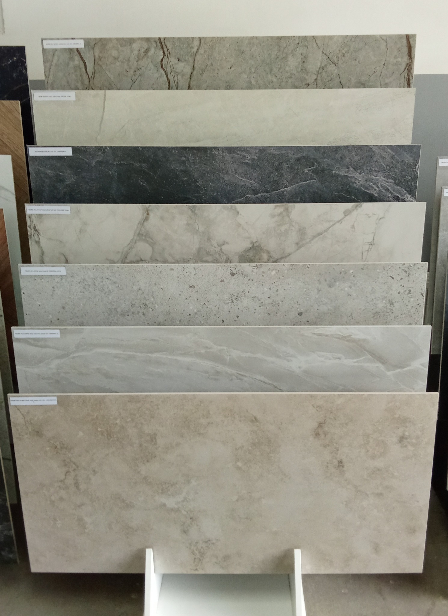 Ekspozycja płytek ceramicznych w różnych wzorach i kolorach, ułożonych warstwowo na białym stojaku. Widoczne etykiety na płytkach, różne faktury i odcienie.