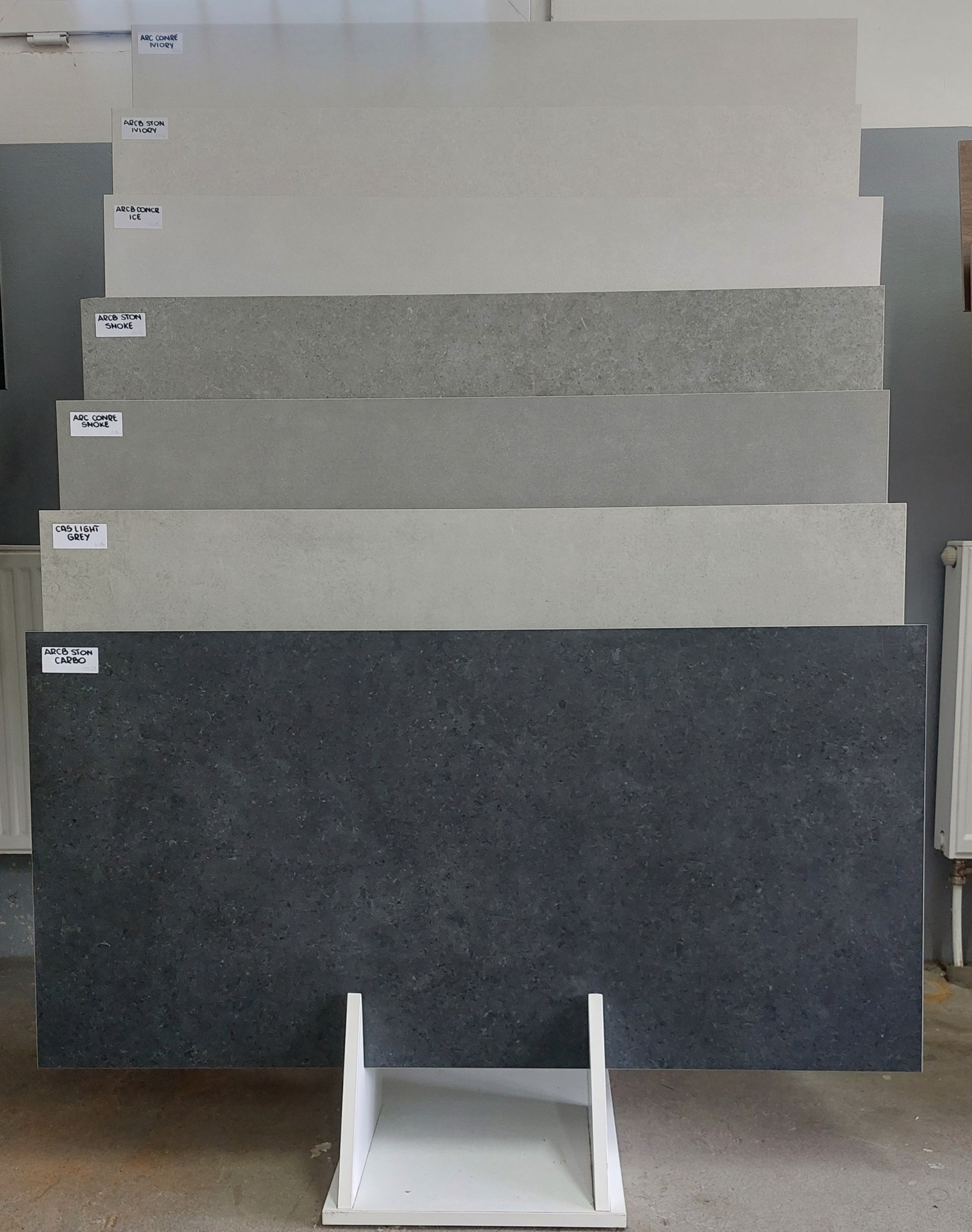 Ekspozycja płytek ceramicznych w różnych odcieniach szarości i bieli na białym stojaku. Prezentacja wzornictwa i faktur płytek.