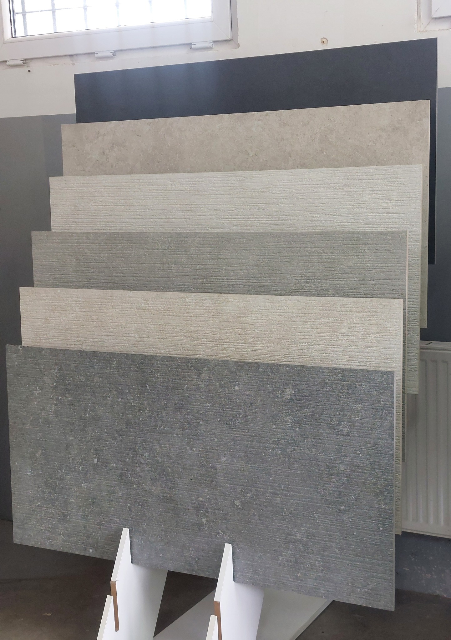 Ekspozycja różnorodnych płytek podłogowych w odcieniach szarości i beżu, prezentowanych na stojaku w jasnym pomieszczeniu z oknem.