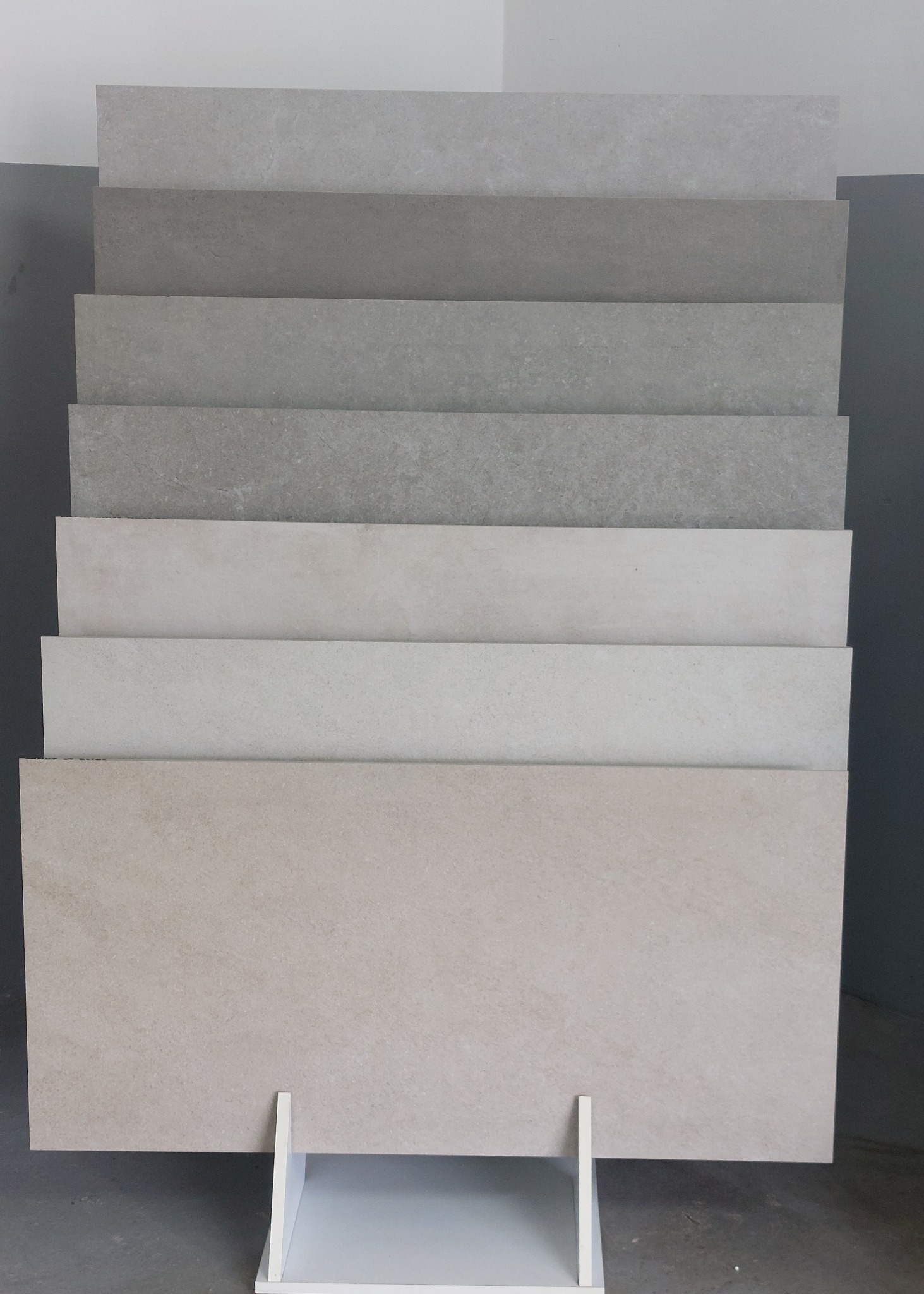 Ekspozycja płytek podłogowych w różnych odcieniach szarości i beżu na białym stojaku. Płytki ułożone warstwowo, od najciemniejszej u góry do najjaśniejszej na dole.
