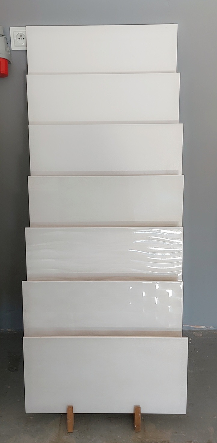 Ułożone pionowo białe płytki ceramiczne o różnych wykończeniach (mat, połysk, satyna) na ekspozycji, widoczne gniazdko elektryczne i fragment instalacji.