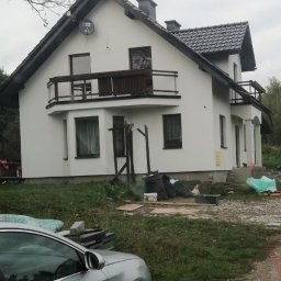 IVAN KUZMYN GREINPLAST BUD - Biały dom z balkonami po tynkowaniu, widoczne narzędzia i worki, częściowo zasłonięty przez srebrny samochód, w tle zieleń.