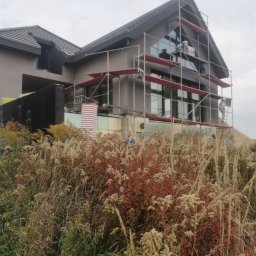 IVAN KUZMYN GREINPLAST BUD - Dom jednorodzinny w trakcie prac elewacyjnych. Widoczne rusztowania, ocieplenie i częściowo pomalowana elewacja. Na pierwszym planie dzika roślinność.