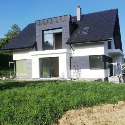 IVAN KUZMYN GREINPLAST BUD - Nowoczesny dom jednorodzinny z szarą elewacją i grafitowym dachem, widoczny z poziomu trawnika. Na elewacji widoczne rusztowania i zabezpieczenia okien.