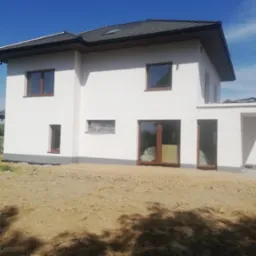 Biała elewacja dwukondygnacyjnego domu z brązowymi oknami, częściowo zabezpieczonymi folią. Widoczny cokół i zagruntowany teren wokół budynku.