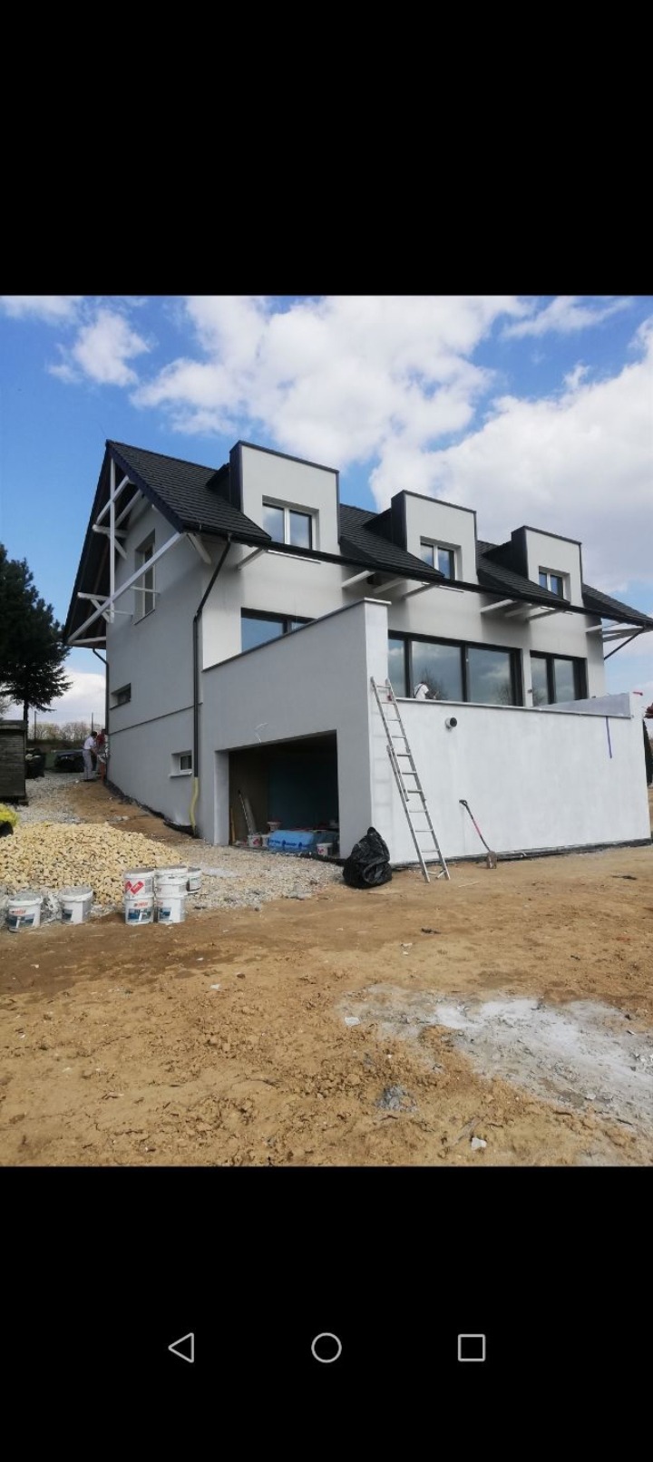 Dwukondygnacyjny dom z białymi ścianami w trakcie prac elewacyjnych. Widoczne wiadra z farbą, drabina oparta o ścianę i pracownicy przy elewacji.