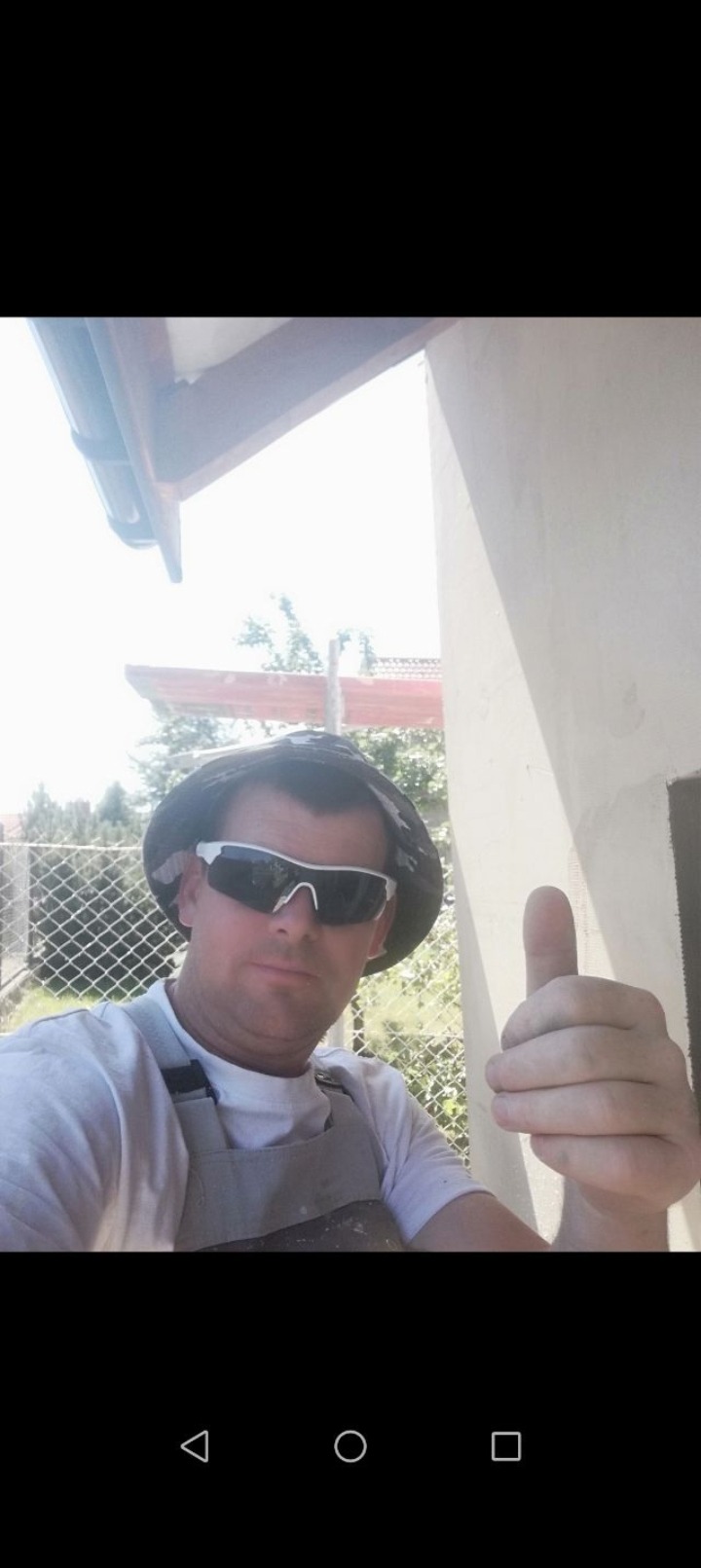 Pracownik w okularach i kapeluszu daje kciuk w górę na tle otynkowanej ściany zewnętrznej domu. Widoczny fragment elewacji i ogrodzenie.