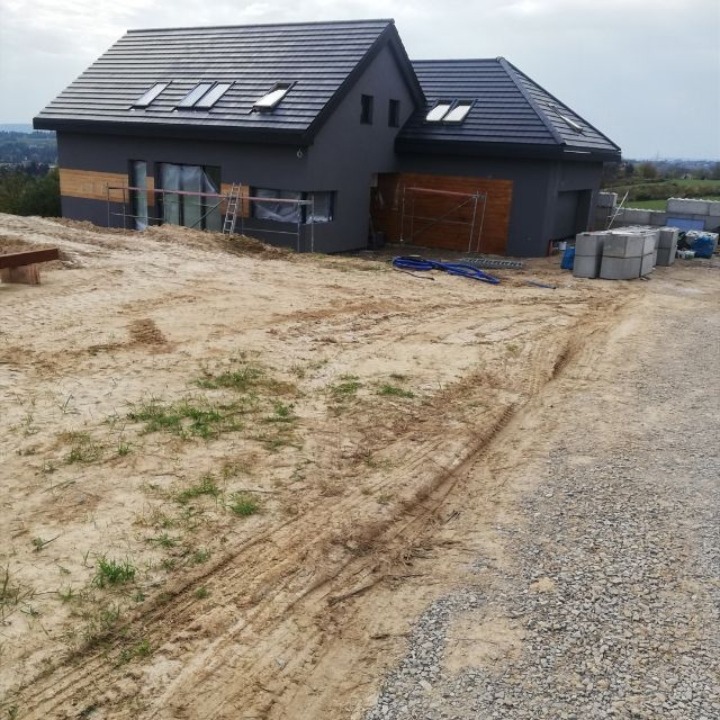 Nowoczesny dom z ciemną elewacją i drewnianymi akcentami, częściowo w rusztowaniach. Widoczne bloczki betonowe i teren w trakcie zagospodarowania.