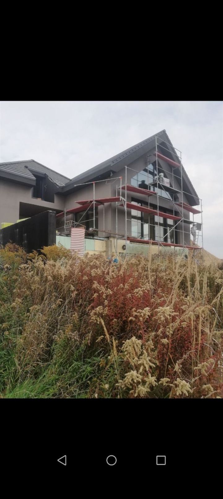 Dom jednorodzinny w trakcie prac elewacyjnych. Widoczne rusztowania, ocieplenie i częściowo pomalowana elewacja. Na pierwszym planie dzika roślinność.