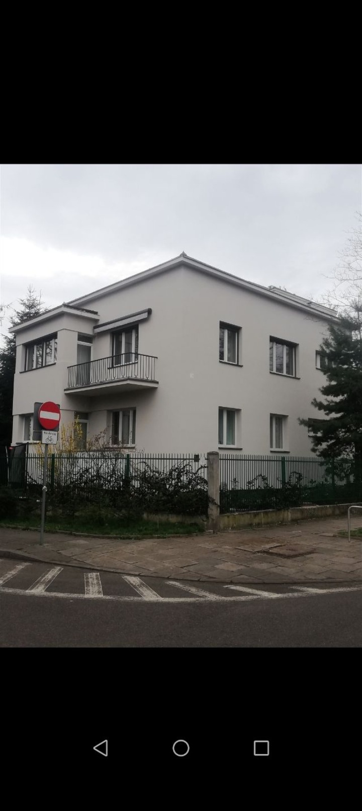 Dwupiętrowy budynek z balkonem, pomalowany na jasny kolor, otoczony zielonym żywopłotem i znakiem drogowym zakazu wjazdu, widziany z ulicy w pochmurny dzień.