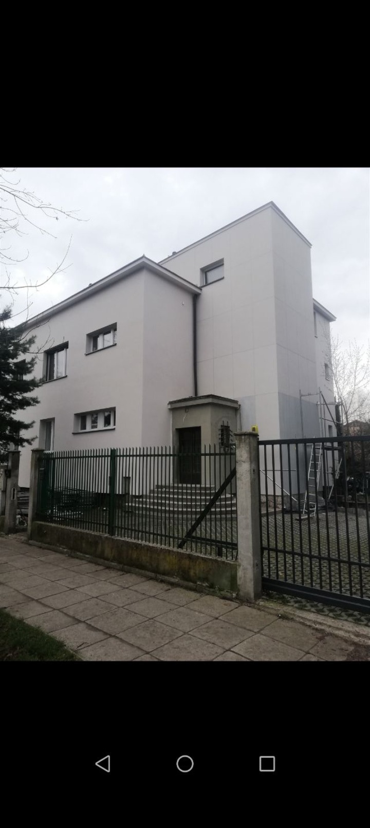 Biały, dwupiętrowy budynek po renowacji elewacji, z widocznymi schodami wejściowymi i czarnym ogrodzeniem. W tle drzewa i pochmurne niebo. Część elewacji jeszcze niepomalowana.