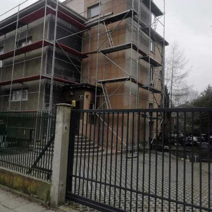 Budynek w trakcie ocieplania, widoczne rusztowania i płyty izolacyjne. Fasadę częściowo pokrywa czarna membrana. Ujęcie zza metalowego ogrodzenia.